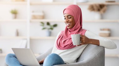 Hafta sonu eğlencesi. Genç siyahi kadın başörtüsü içinde evde dizüstü bilgisayar ve kahveyle dinleniyor, başörtülü Afrikalı Müslüman kadın internette sörf yapıyor ve sıcak içeceğin tadını çıkarıyor, boşluğu kopyalıyor.