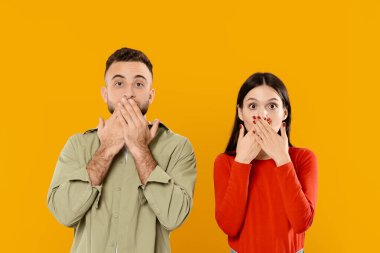 Genç bir çift, ağızlarını ellerle kapatarak neşeli bir sürpriz sergiliyor. Parlak sarı bir zemin üzerine kurulmuş canlı ifadeleri ilişkilerinde neşe ve şefkat taşır..