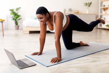 Plump young Afro woman working out butt muscles in front of laptop, following video tutorial, making exercises for weight loss at home. Overweight black lady training indoors during covid quarantine