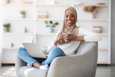 Yurt içi eğlence. Neşeli siyah Müslüman kadın evde dizüstü bilgisayar ve kahveyle dinleniyor, mutlu İslami kadın internette geziniyor ve oturma odasında dinlenirken sıcak içkinin tadını çıkarıyor.