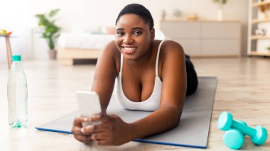 Elinde bir şişe su ve akıllı telefon olan şişman siyahi kadın internette geziniyor, evdeki sosyal ağları kontrol ediyor. Fazla kilolu Afrikalı Amerikalı kadın cep telefonu kullanarak ev içi sporlara ara veriyor.