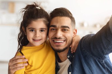Orta Doğulu mutlu bir babanın, küçük kızıyla evde selfie çekerken, neşeli genç bir Arap baba ve sevimli küçük bir kızın kameraya sarılıp gülümsediği fotoğraf.