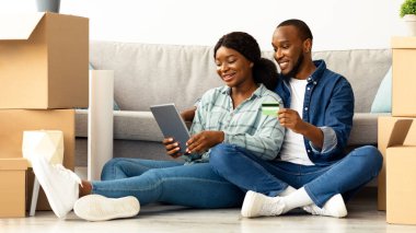 Yeni dairelerine taşındıktan sonra dijital tabletle ve kredi kartıyla gülümseyen genç siyah eşler internetten mutlu Afrikalı Amerikalı çiftler internetten alışveriş yapıyorlar.