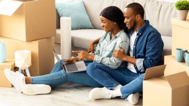 Çevrimiçi sipariş. Genç siyah çift dizüstü bilgisayar kullanıyor, yeni dairelerine taşındıktan sonra internetten mobilya alıyorlar, mutlu Afrika kökenli Amerikalı eşler ellerinde bilgisayarla karton kutuların arasında oturuyorlar.