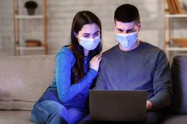 Coronacrisis Concept. Cerrahi maskeli ciddi bir çift haber okuyor ve kanepede oturup bilgisayar izliyor.