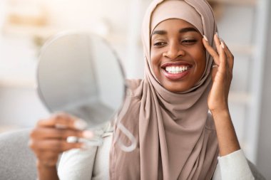 Güzellik Konsepti. Başörtüsü içindeki çekici siyahi kadın büyüteç aynasına bakıyor, başörtülü mutlu Afrikalı Müslüman kadın yumuşak tenine dokunuyor ve gülümsüyor, yakın çekim