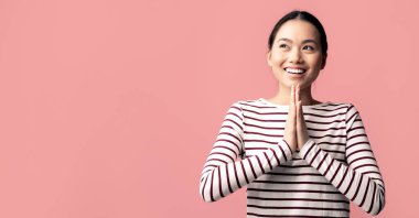 Asyalı genç, rüya gibi bir bayan elleri bağlı ve yana bakıyor olumlu, Koreli bir kadın bir şey diliyor ya da merak ediyor pembe arka planda tek başına duruyor ve uzayı kopyalıyor.