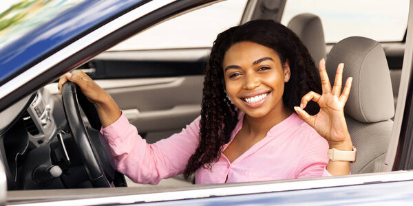 Великолепный автомобиль. Счастливая афроамериканка демонстрирует хорошо знак Жест, сидя в новом автомобиле покупая современный автомобиль в магазине дилеров, наслаждаясь отпуска, глядя на камеру через окно