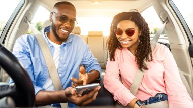 Yaz Tatili Konsepti. Lüks arabanın içinde oturan gülümseyen Afrikalı çiftin portresi, cep telefonunu gösteren neşeli adam, dijital harita uygulaması kullanarak yerini gösteriyor.