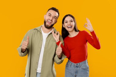 Genç bir çift sevgisini geniş bir şekilde gülümseyerek ve birlikte şakacı jestler yaparak gösterir. Sıradan giyiniyorlar, parlak sarı bir arka plana karşı canlı bir bağlantı sergiliyorlar..