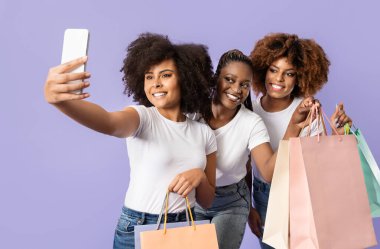 Üç genç Afrikalı Amerikalı kadın alışveriş torbaları tutarken selfie çekerken mutlu bir şekilde gülümsüyor. Sıradan giyinirler ve pastel arka planda birlikte vakit geçirmenin tadını çıkarırlar..