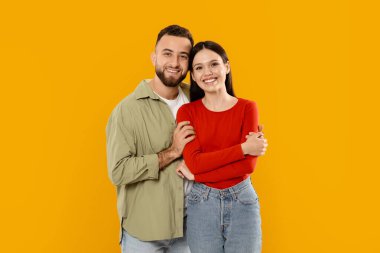 Genç bir çift birbirine yakın duruyor, kameraya samimi bir şekilde gülümsüyor. Sevgi ve mutluluk gösterirler. Parlak sarı bir arkaplan önünde dururlar. Aşkı ve birlikteliği tasvir ederler..