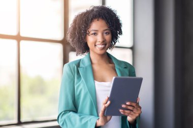 Parlak bir ofis ortamında, kıvırcık saçlı genç bir profesyonel gülümserken bir tablet tutar. Bir pencerenin yanında duruyor, takım çalışmasını ve şirket dünyasındaki yenilikleri yansıtıyor..