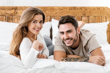 Mutlu milenyum çifti covid-19 karantinası sırasında kendilerini izole ediyorlar. Gülümseyen genç adam ve kadın tahta yatakta uzanır, gün ışığında kameraya bakar yatak odasının kırsal kesiminde, panorama
