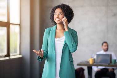 Yeşil ceketli bir kadın akıllı telefonuyla konuşuyor, modern bir şirket ofisinde fikirlerini güvenle dile getiriyor. Bir meslektaşım arka planda bir masada çalışıyor, takım çalışmasını vurguluyor..