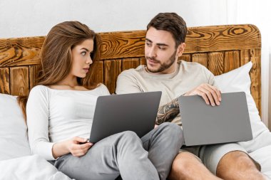 Modern akşam eğlencesi. Gündelik giysiler içindeki erkek ve kız dizüstü bilgisayarlarla yatağa uzanıyor ve yatak odası, panorama, boş yer içinde konuşuyorlar.