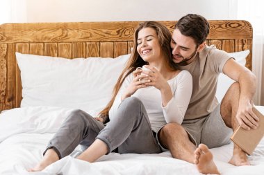 Ev konseptinde kal. Kitaplı adam, yatakta fincanlı kız.