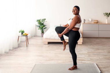 Şişman siyahi kadın bacağını esnetiyor yoga minderinin üzerinde esneklik egzersizleri yapıyor evdeki yoğun kilit altında aktif bir yaşam tarzı sürüyor. Artı, içeride çalışan Afrikalı bir kadın, boş yer.