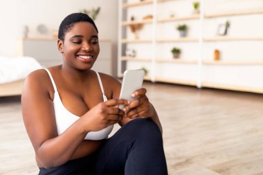 Gezici cihaz kullanan kıvrımlı siyah kadın, ev içi eğitimden arayı alıyor, kapalı alanda. Tombul Afrikalı Amerikalı kadın sosyal medyada geziniyor, ev çalışmasından sonra dinleniyor.