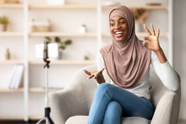 Evde video kaydetmek için Smartphone kullanan mutlu siyah Müslüman kadın blogcu, video günlüğü için içerik yakalıyor, konuşuyor ve cihaz kamerasının önünde el kol hareketi yapıyor.
