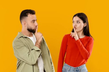 Genç bir çift canlı sarı bir arkaplanın önünde birlikte duruyor. Sessizliğin neşeli bir anını parmak dudağa hareketleriyle gösteriyorlar. Bağlarını ve sevgilerini yansıtıyorlar..