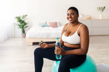 Artı, spor salonunda elinde bir şişe suyla oturan boyunda bir zenci kadın tecrit sırasında evde egzersiz yapıyor. Aşırı kilolu Afrikalı Amerikalı kadın iç egzersizler sırasında susuz kalmıyor.