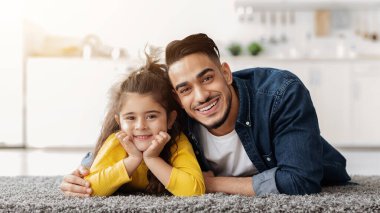 Babanla vakit geçir. Şirin küçük kız ve Arap babasının evde poz verip yumuşak halıda rahatladıkları, neşeli bir Orta Doğulu baba ve çocuğun kameraya bakıp gülümsedikleri ve boş bir alan aradıkları portresi.