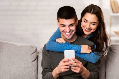 Milenyum çifti cep telefonlarını paylaşıyor evlerindeki koltukta oturuyor, kız erkeğine arkadan sarılıyor.