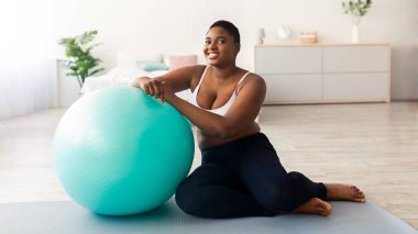 Tombul siyah bayan fitness topunun yanında oturuyor, evde spor minderiyle çalışıyor. Aşırı kilolu Afrikalı Amerikalı kadın kapalı alanda ev eğitimi alıyor, tam boy.