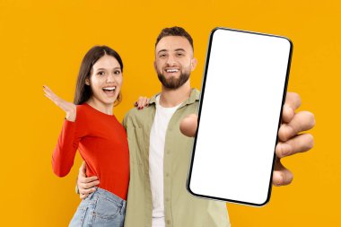 Genç beyaz bir çift ellerinde akıllı bir telefonla hevesle poz veriyor. Kadın büyük bir gülümsemeyle gülümserken, adam sevgisini ve sevgisini göstererek mutlu bir şekilde sırıtıyor..