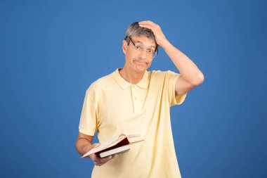 A middle aged Caucasian man in a yellow shirt appears confused while holding an open book. The plain blue background emphasizes his expression of uncertainty and contemplation. clipart