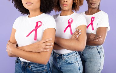 Three young African American women stand together against a purple backdrop, arms crossed. They wear white shirts with pink ribbons, showcasing their support for breast cancer awareness. clipart