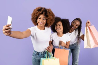 Üç genç Afrikalı Amerikalı kadın gülümsüyor ve renkli alışveriş torbaları tutarken selfie için poz veriyorlar. Canlı bir ortamda dostluk ve neşe sergiliyorlar, günlerini kutluyorlar..