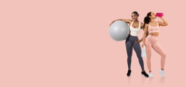 İki kadın spor salonunda aktif bir antreman yapıyor. Biri büyük bir egzersiz topu tutarken, diğeri bir spor şişesinden içerek sağlıklı bir yaşam tarzı sergiliyor..