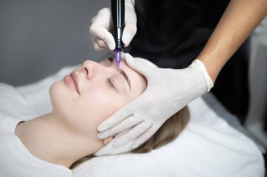 Bir müşteri modern bir kuaförde dinlenir ve bir teknisyen kaşlarını büyütmek için dikkatlice kalıcı makyaj yapar. Mekan, uzman hizmetleri yoluyla güzelliği ve şımartmayı teşvik ediyor..