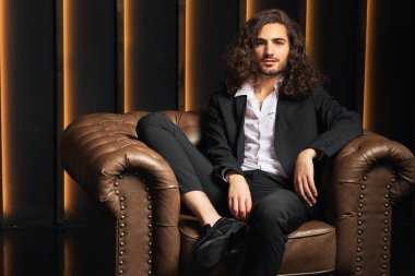 Uzun, kıvırcık saçlı, iyi giyimli genç bir adam deri bir koltukta rahat rahat oturur ve modern ofis ortamında kendine güveni ve tarzı ile sıcak bir ışık saçar..