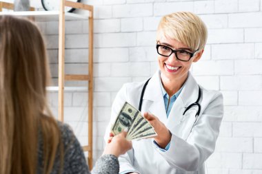 Yolsuzluk konseri. Gülümseyen kadın doktor ofisin içindeki hastaların ellerinden para alıyor.