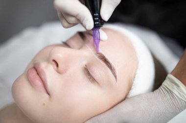 Bir kadın bir güzellik salonunda kaşlarına titiz bir kalıcı makyaj prosedürü uygulanır. Teknisyen doğal bir görünüm sağlamak için renkleri titizlikle uyguluyor..