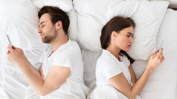 Marital Crisis, Indifference. Couple Using Cellphones Lying Back-To-Back In Bed Indifferent To Each Other. Millennial Spouses Ignoring One Another Texting With Lovers At Home. View From Above