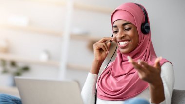 Mesafe iletişimi. Tesettürlü neşeli siyahi kadın ve kulaklık evde dizüstü bilgisayarla video görüşmesi yapıyor, Olumlu Afrikalı Müslüman kadın çevrimiçi toplantı ve internet konferansından hoşlanıyor,