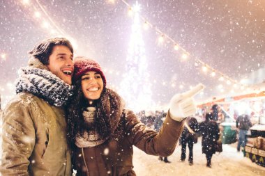 Xmas. Neşeli kız arkadaş bir şey gösteriyor erkek arkadaşı parmağıyla işaret ediyor geceleri dışarıda Noel pazarında eğleniyor.