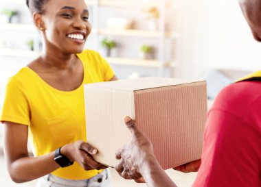 Paket kutusunu teslim eden teslimatçı çocuk evinin kapısında duran mutlu Afrikalı Amerikalı kadına teslim ediyor. Bayan Müşteri Kırmızı Üniformalı Erkek Kurye 'den karton paket alıyor. Seçici Odaklanma