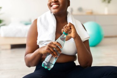 Evinde temiz su şişesiyle spor minderinde oturan, tanınmayacak kadar kilolu, genç siyahi bir bayan, yakın plan. Ayrıca ev içi çalışma sırasında susuz kalmayan Afrikalı Amerikalı bir kadın.