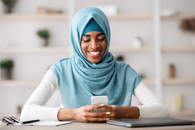 Ofiste tesettürde gülümseyen Afrikalı İslami Kadın portresi, cep telefonuyla masada oturan, internete göz gezdiren, internetten alışveriş yapan veya mesajlaşan neşeli siyahi Müslüman kadın.