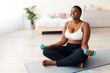 Spor kıyafetli şişman siyahi bir kadın yoga minderinde oturuyor ve evde dambıllarla egzersiz yapıyor. Milenyum Afrikalı Amerikalı kadın iç güç egzersizi yapıyor.