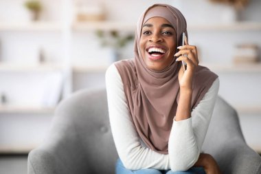 Tesettürlü neşeli siyah Müslüman kadın evde cep telefonuyla konuşuyor, oturma odasında rahat koltukta dinleniyor, mutlu dindar Afrikalı kadın arkadaşıyla hoş sohbetler yapıyor, boşluğu kopyalıyor.
