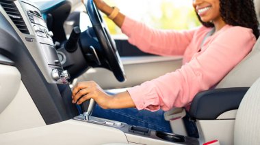 Kadın şöför. Kentte lüks otomobil sürmeyi öğrenen gülümseyen Afrikalı kadın portresi sürücü koltuğunda oturuyor, direksiyon tutuyor ve vites koluna bakıyor.