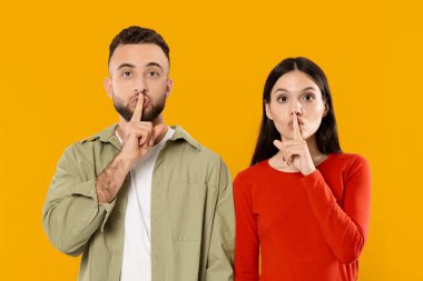 Genç evli bir çift, parmakları dudaklarında poz verir, neşeli bir gizlilik jesti ya da parlak sarı bir arka plana karşı sessizlik gösterirler, sevgilerini gösterirler..