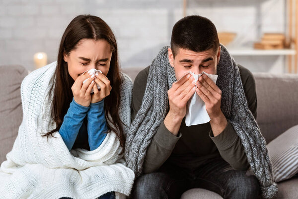 Sick young couple blowing noses sitting on couch, having flu together, copy space