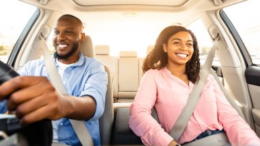Modern lüks araba kullanan, gülümseyen, Afro-Amerikan bir adamın ön manzara portresi. Ön koltukta oturan mutlu, güzel, genç bir kadın, yola bakıyor, parlıyor.